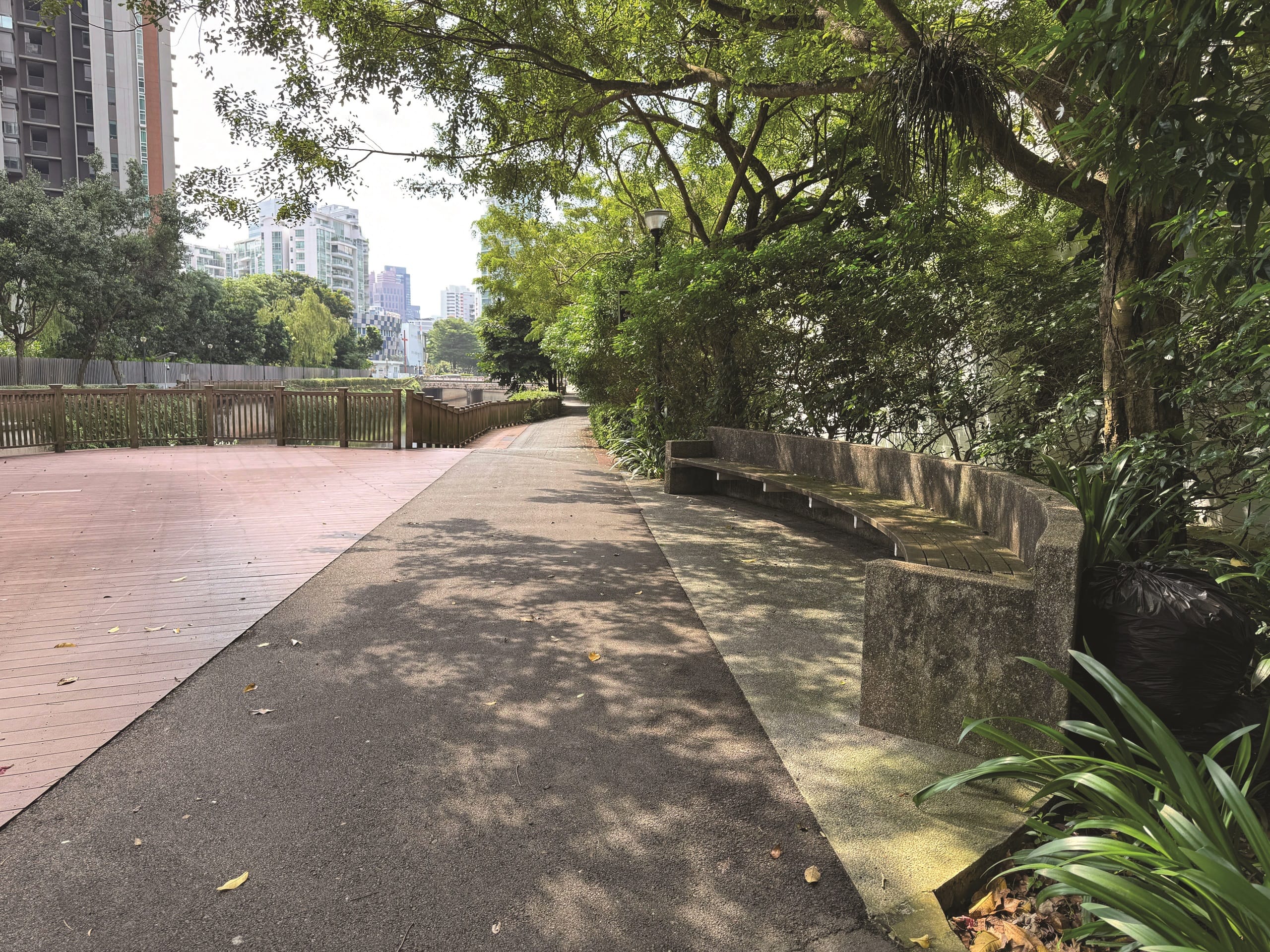 A park path with trees and buildings in the distance. A curved stone bench sits beside the path.